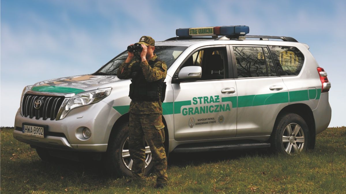 Funkcjonariusz Straży Granicznej patrzy przez lornetkę na tle samochodu oznakowanego "Straż Graniczna"