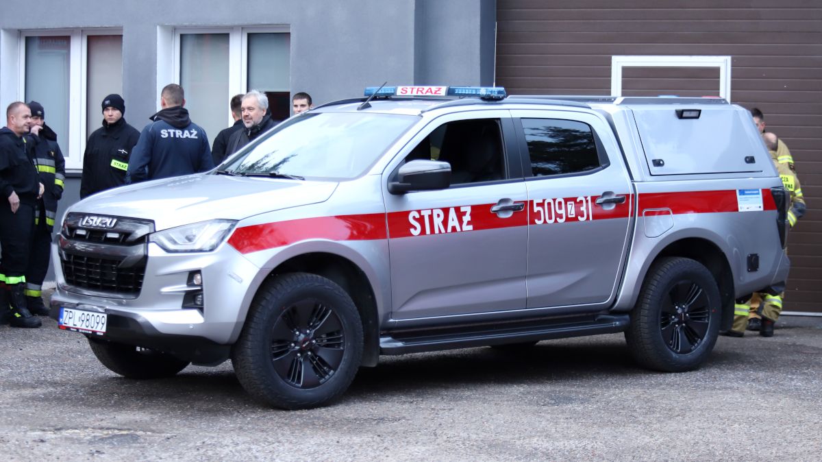Na pierwszym planie lekki samochód rozpoznawczo-ratowniczy. w tle osoby i budynek remizy w Tanowie