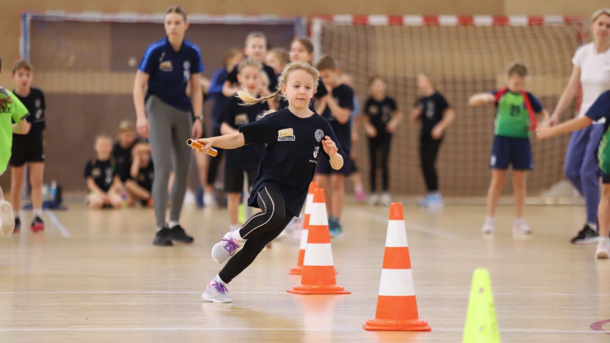 Dziewczynka biegnie z pałeczką, omijając pachołki, w tle inni uczestnicy zawodów