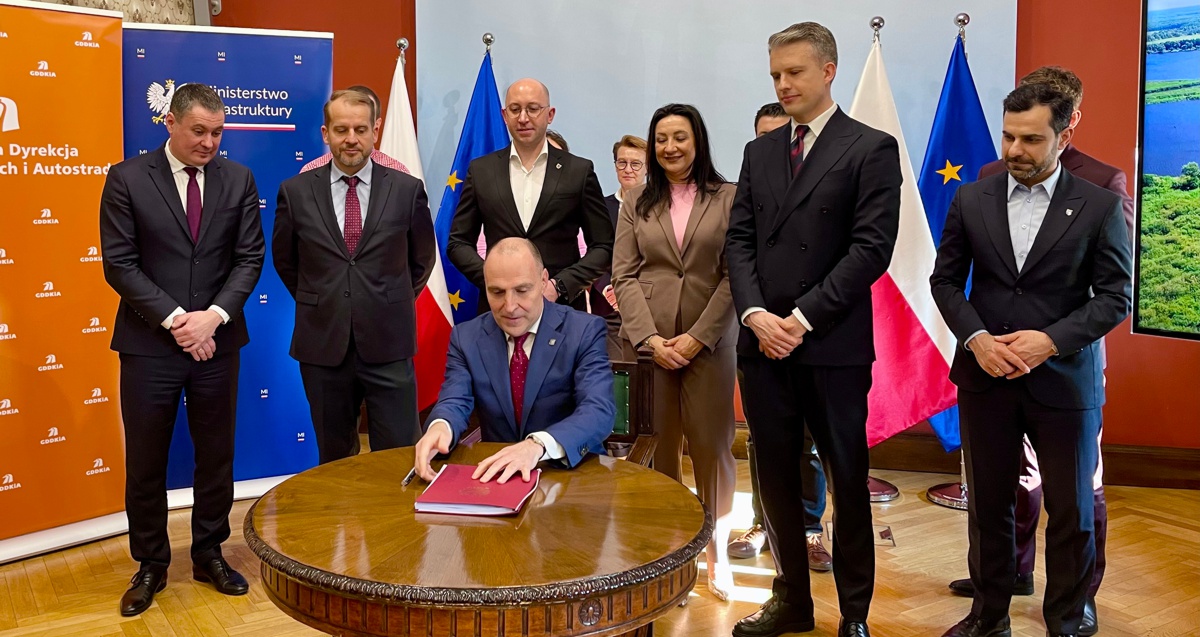 Wojewoda siedzi przy stole i podpisuje dokument. Z ty za nim stoją przedstawiciele ministerstwa, samorządów i firm.