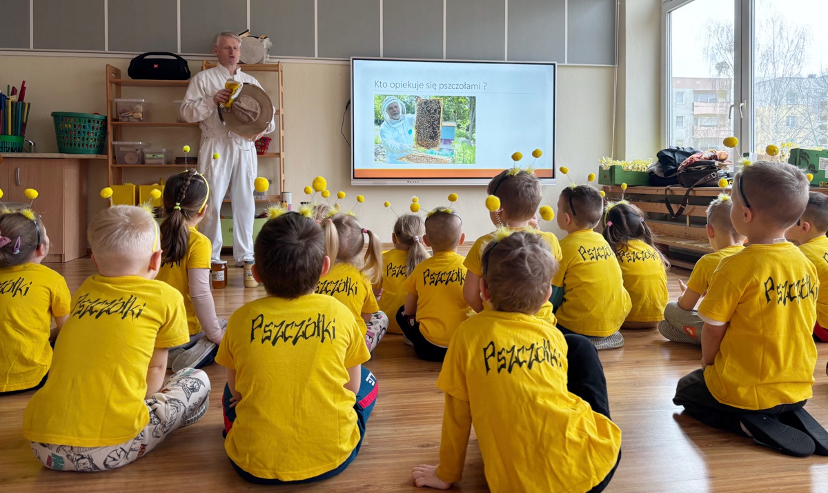 Na pierwszym planie dzieci siedzą tyłem na podłodze i słuchają mężczyzny ubranego w strój pszczelarza
