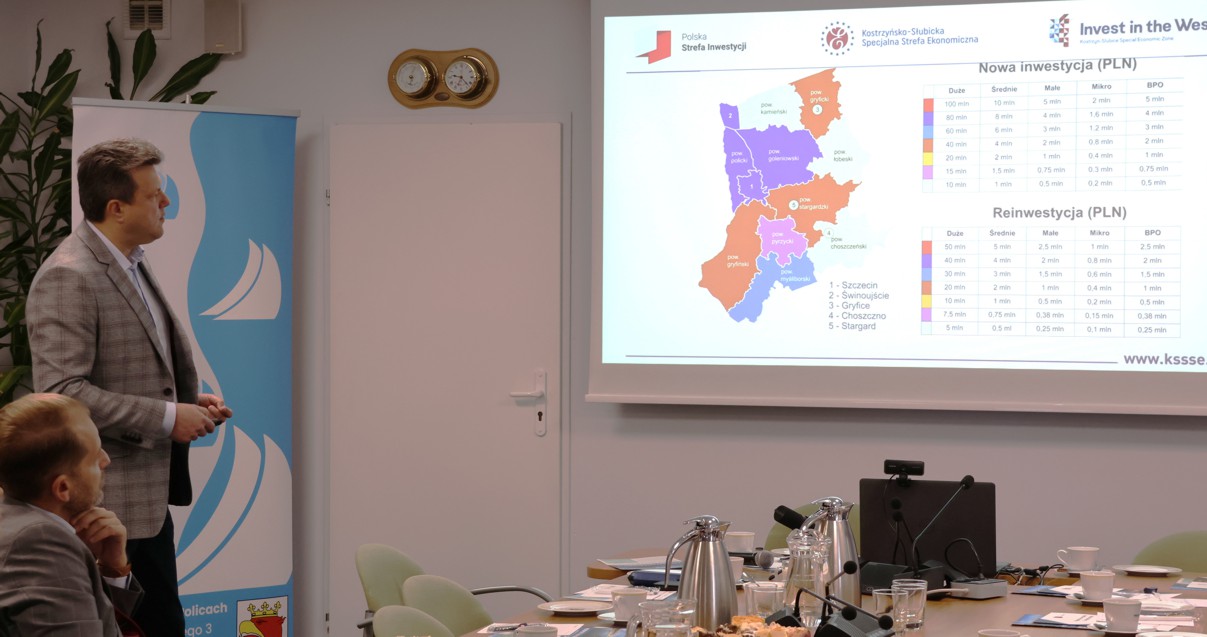 Dwie osoby jedna stoi, druga siedzi a w tle dane wyświetlane z rzutnika