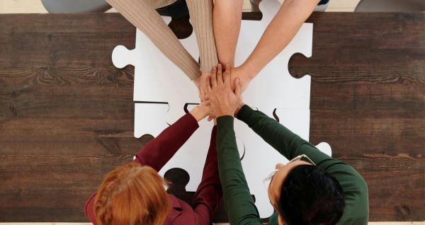 Zdjęcie zrobione z góry czterech osób, które złączyły ręce na środku nad stołem na  którym leżą duże puzzle