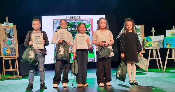 Piątka nagrodzonych dzieci stoi na scenie trzymając worki z nagrodami, po bokach na sztalugach prace uczestników konkursu