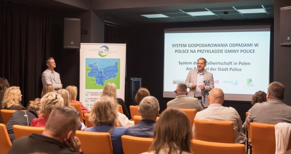 Dwie osoby stoją, jedna na tle planszy druga z boku. Przed nimi siedzą osoby.