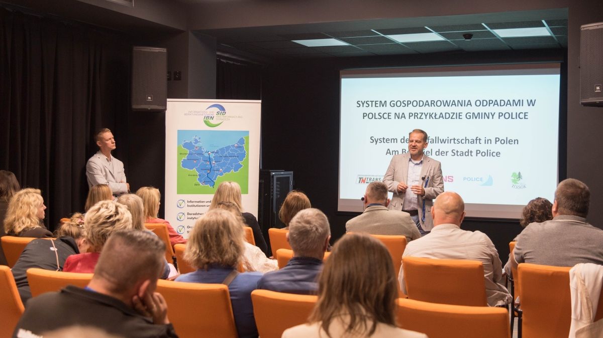Dwie osoby stoją, jedna na tle planszy druga z boku. Przed nimi siedzą osoby.