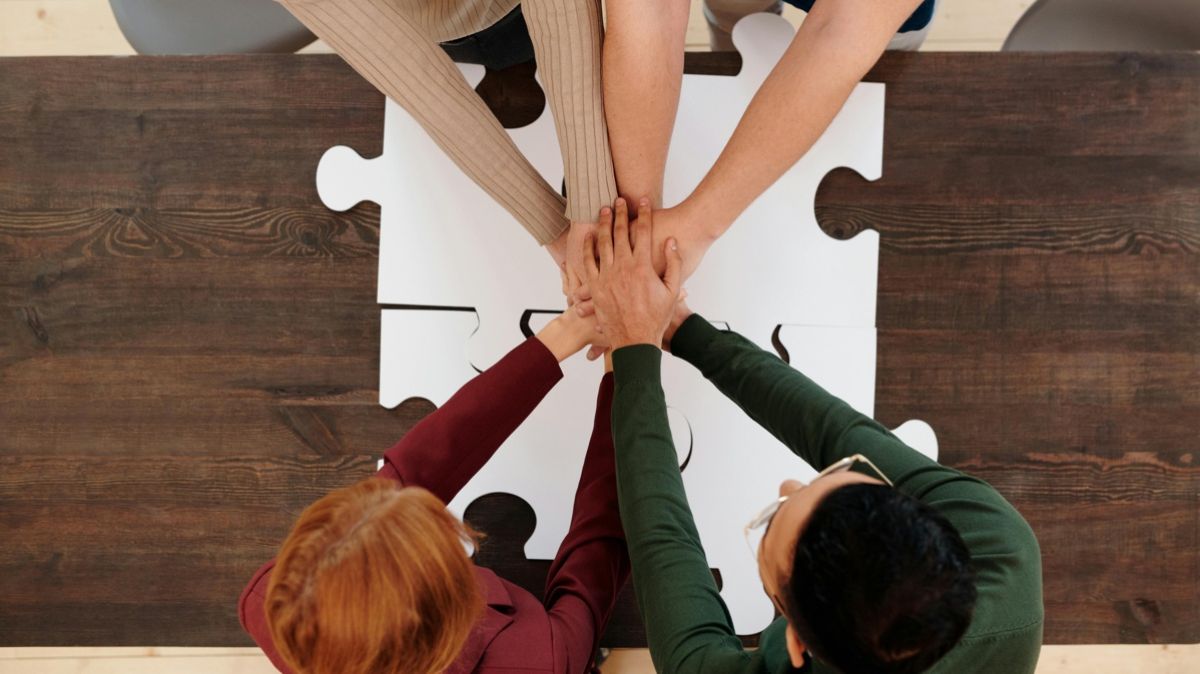 Zdjęcie zrobione z góry czterech osób, które złączyły ręce na środku nad stołem na  którym leżą duże puzzle