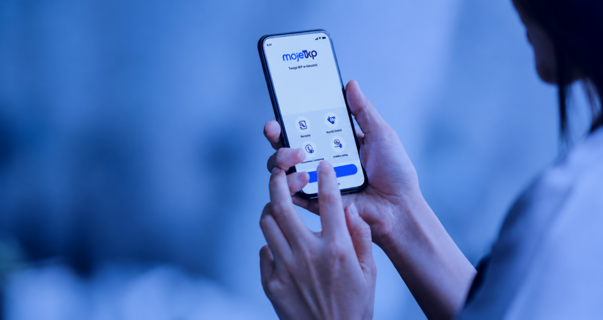 Grafika na której jest częściowo widoczna osoba trzymająca w dłoni telefon, na ekranie którego wyświetla się aplikacja IKP