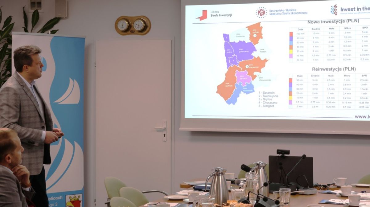 Dwie osoby jedna stoi, druga siedzi a w tle dane wyświetlane z rzutnika