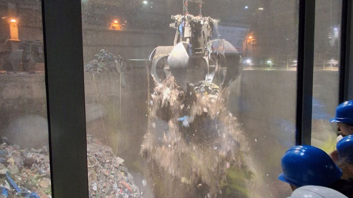 Grupa uczniów w kaskach patrzy przez szybę jak przenoszone są śmieci chwytakiem.