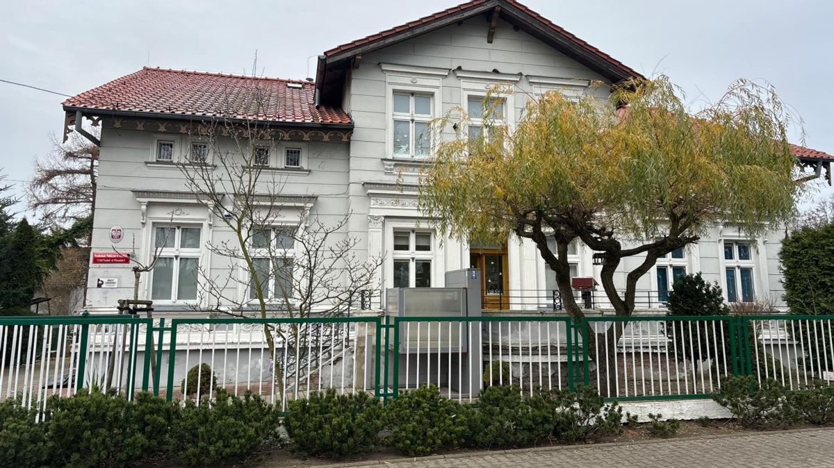 Na pierwszym planie ogrodzenie metalowe, za nim dwa drzewa, w tle budynek przedszkola w Trzebieży