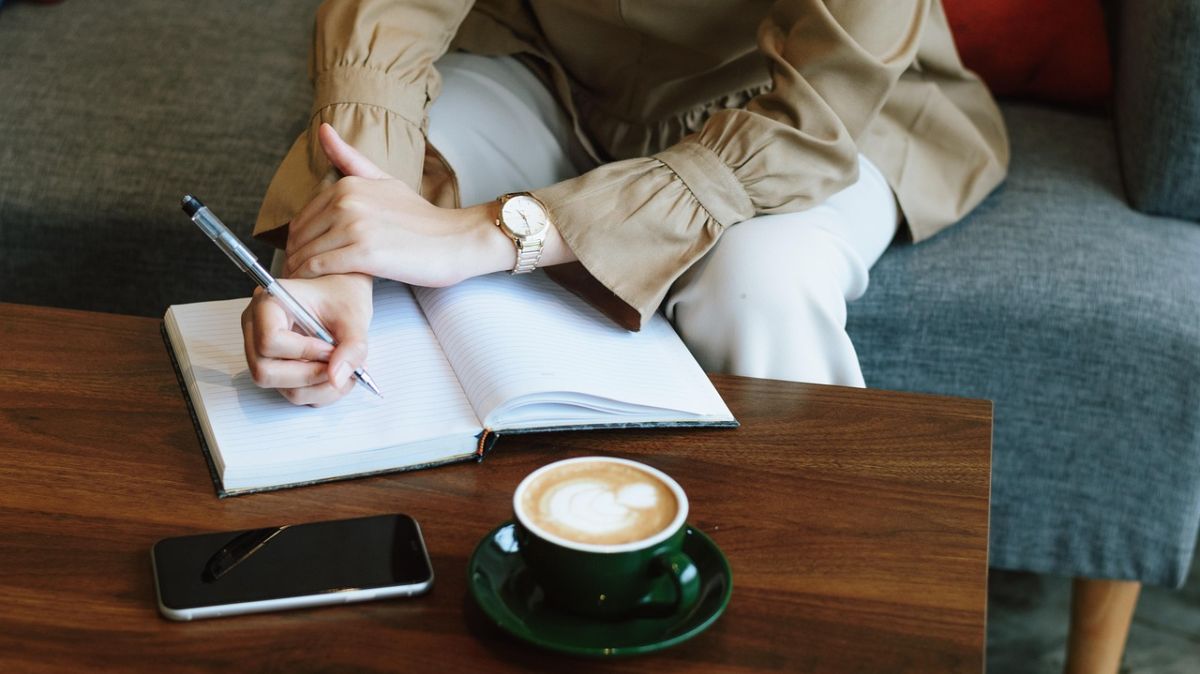 Osoba siedząca przy ławie pisze coś w notesie, na stole stoi kawa
