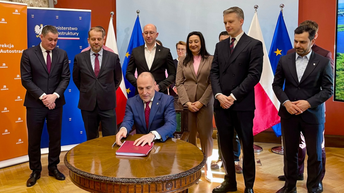 Wojewoda siedzi przy stole i podpisuje dokument. Z ty za nim stoją przedstawiciele ministerstwa, samorządów i firm.