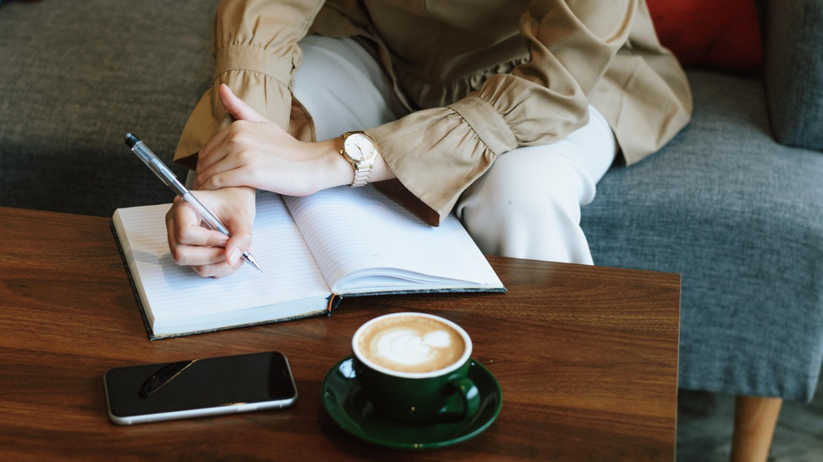 Osoba siedząca przy ławie pisze coś w notesie, na stole stoi kawa