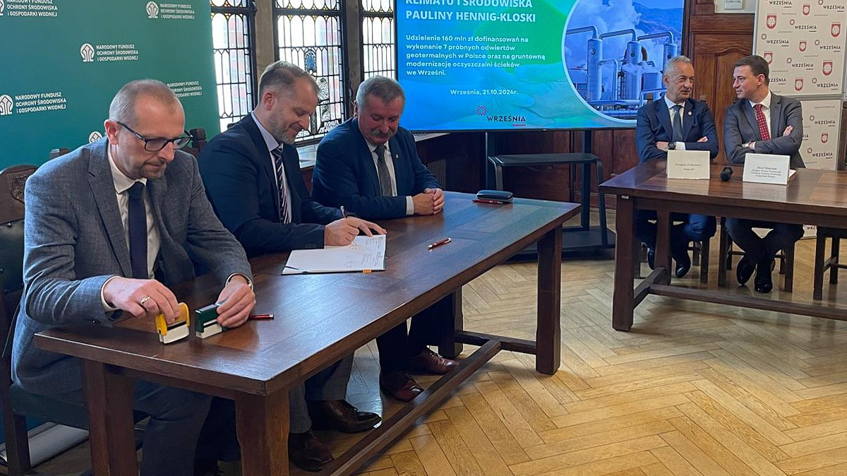 Na pierwszym planie stół a za nim siedzą trzy osoby i podpisują umowę, w głębi część stołu przy którym siedzą dwie osoby