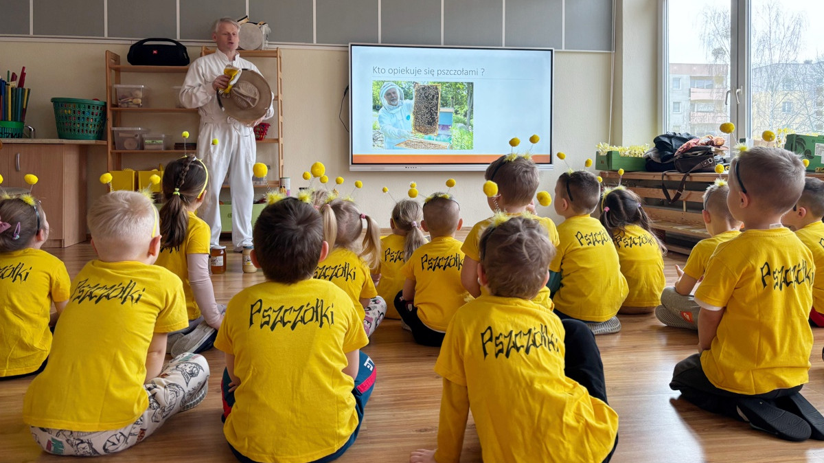 Na pierwszym planie dzieci siedzą tyłem na podłodze i słuchają mężczyzny ubranego w strój pszczelarza
