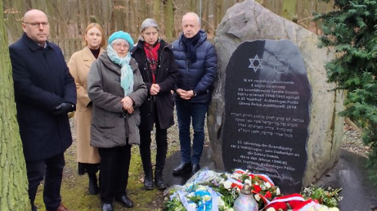 Trzy kobiety, dwóch mężczyzn stoi obok obelisku poświęconego ofiarom filii obozu koncentracyjnego w Stutthofie. Przed obeliskiem leżą wiązanki kwiatów.