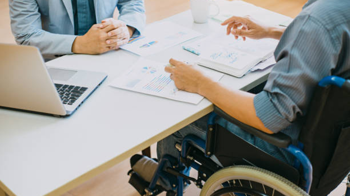 Dwie osoby siedzą przy stole, jedna na wózku inwalidzkim, na stole leżą dokumenty i stoi laptop